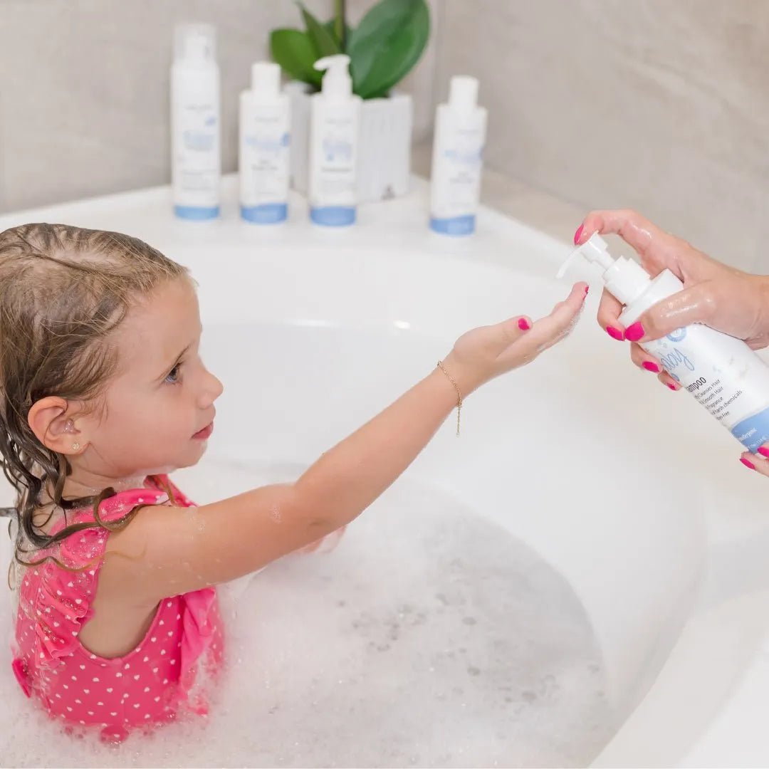 A child in a pink swimsuit reaches for Baby Jolie 2-in-1 hair and body wash dispensed from a white pump bottle in a bubble bath.