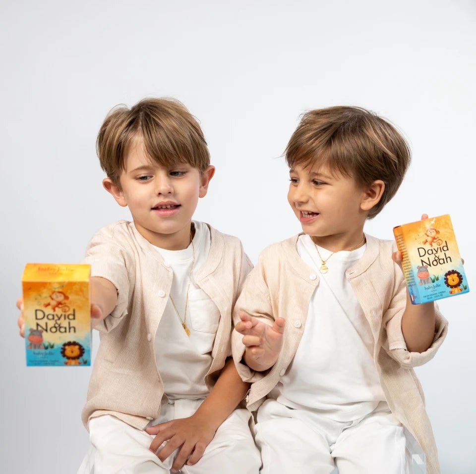 Two young boys in beige outfits holding David & Noah baby body wash and cologne boxes with colorful animal illustrations.