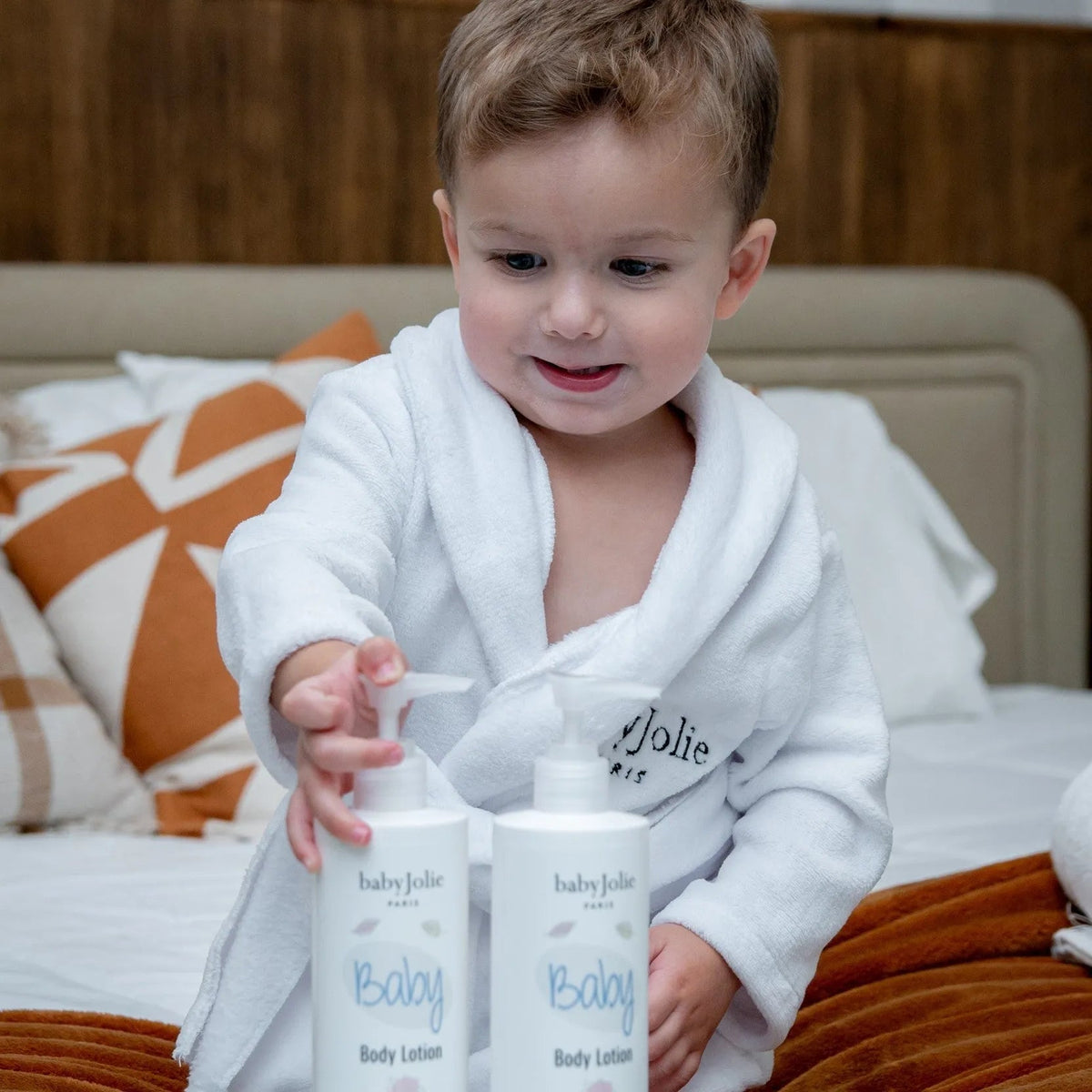 Toddler in a white bathrobe reaching for two 11oz bottles of Baby Jolie body lotion on a bed.