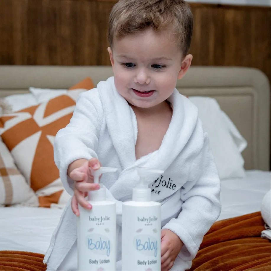 Toddler in a white plush robe reaching for two Baby Jolie body lotion bottles on a bed with brown and white pillows.