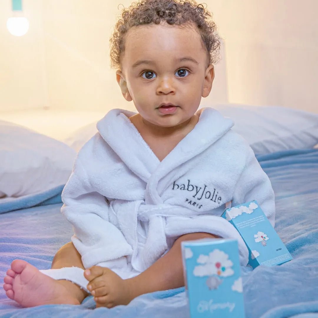 Baby in a white plush robe sitting on a blue blanket next to Baby Jolie lotion and perfume boxes.