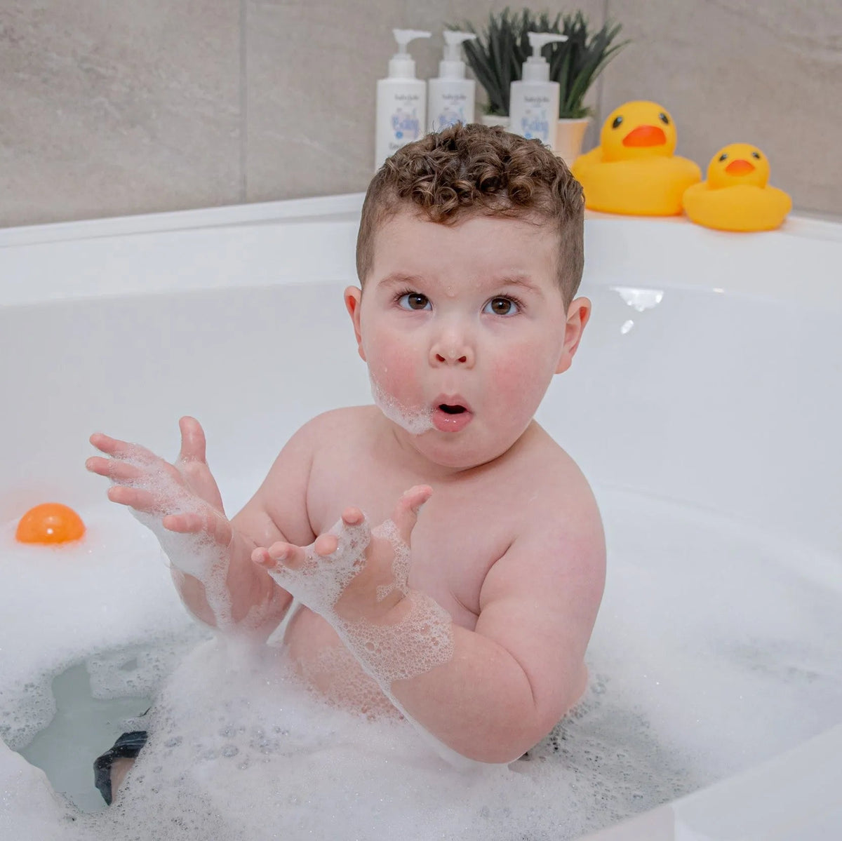 Toddler playing with soapy bubbles in a white bathtub with Baby Jolie shampoo bottles and yellow rubber ducks nearby.
