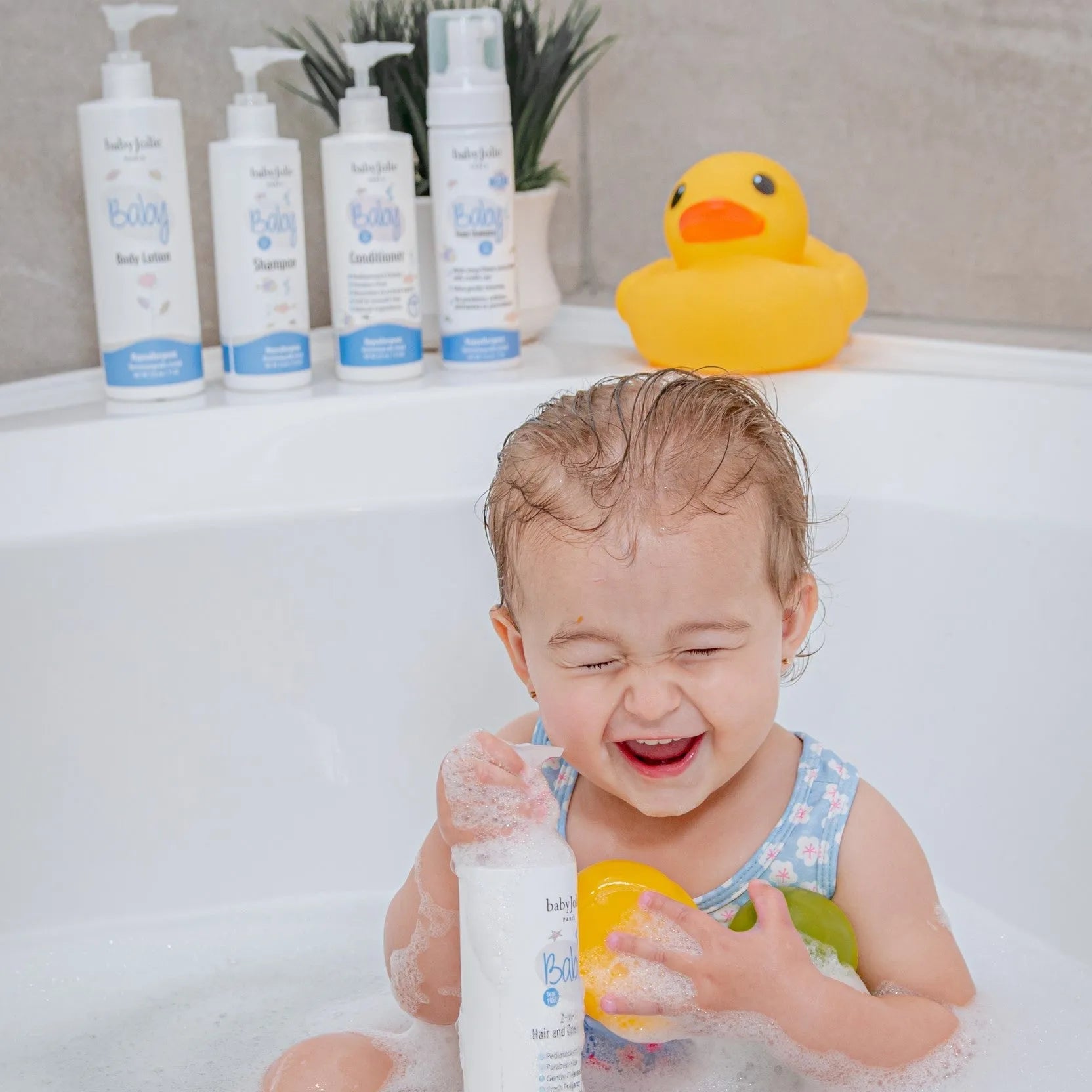 Happy toddler playing with Baby Jolie shampoo bottle in a bubble-filled bathtub with a yellow rubber duck nearby.