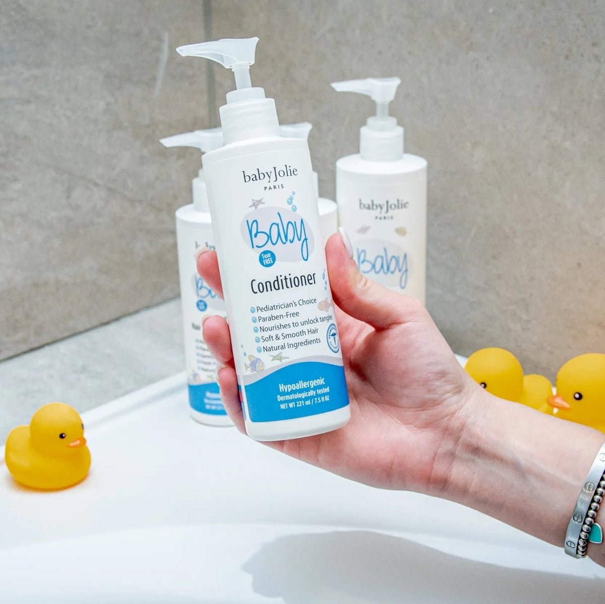Hand holding a Baby Jolie conditioner bottle with a pump dispenser, surrounded by yellow rubber ducks in a bathroom.
