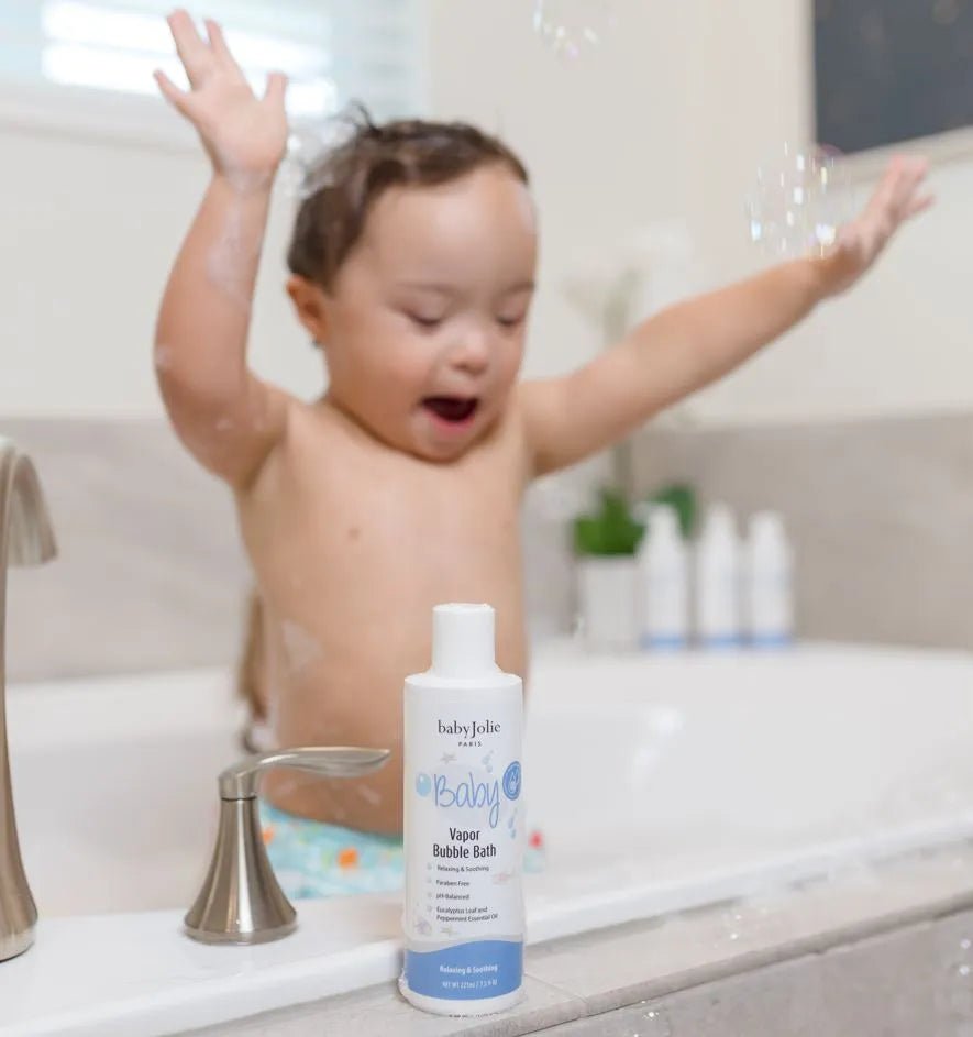 Baby Jolie Vapor Bubble Bath bottle on a tub ledge with a happy toddler playing with bubbles in the background.