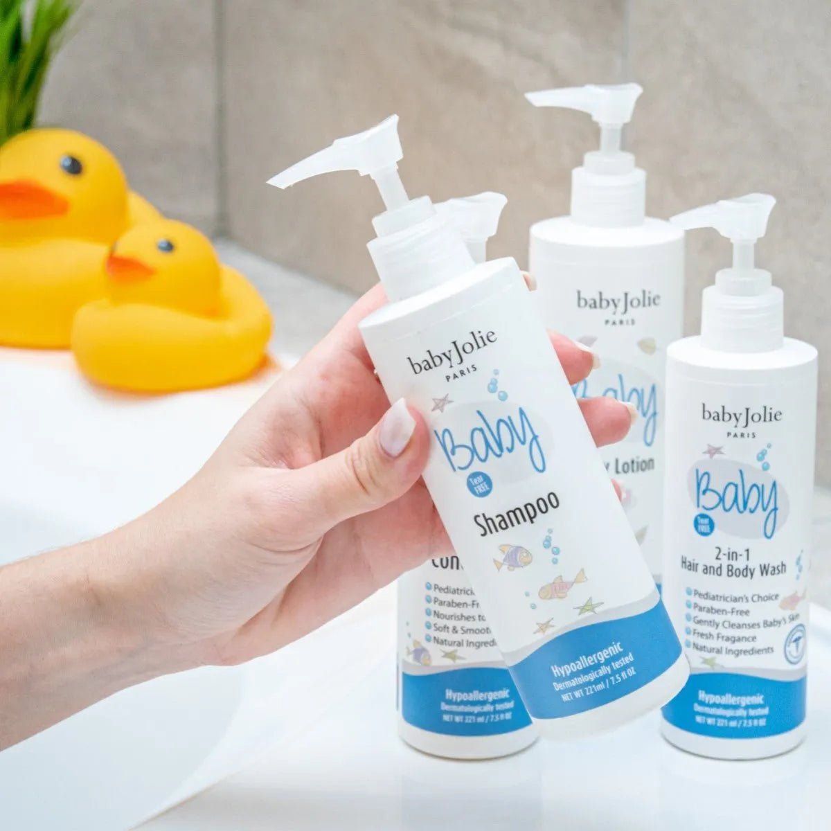 Hand holding a Baby Jolie shampoo bottle with blue accents and fish graphics, with matching bath products and rubber ducks.