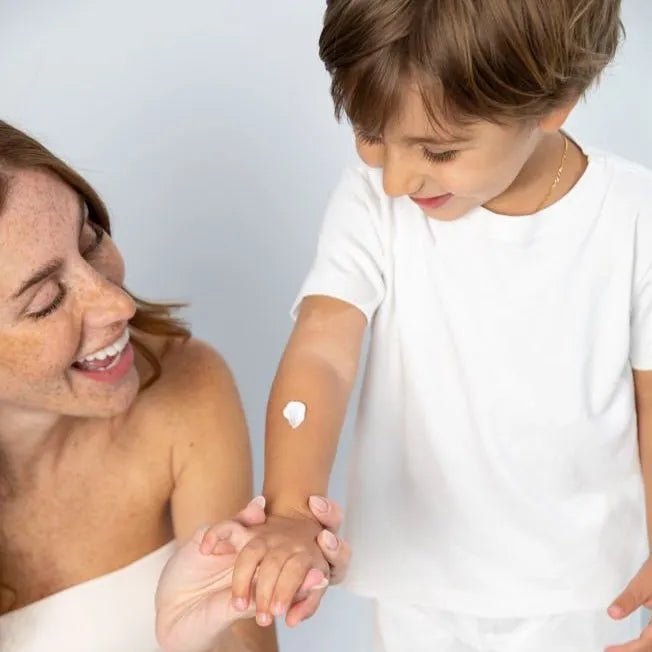 A smiling woman applies a dab of white David & Noah moisturizing body lotion to a young boy's arm.