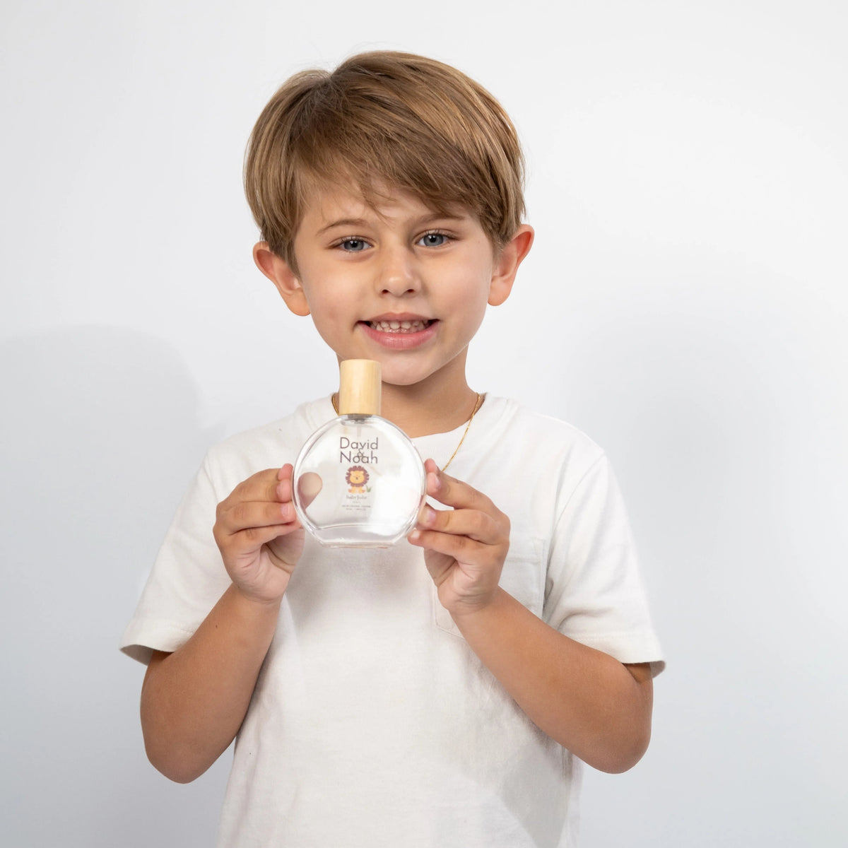 Smiling young boy in a white t-shirt holding a round David & Noah perfume bottle with a wooden cap.