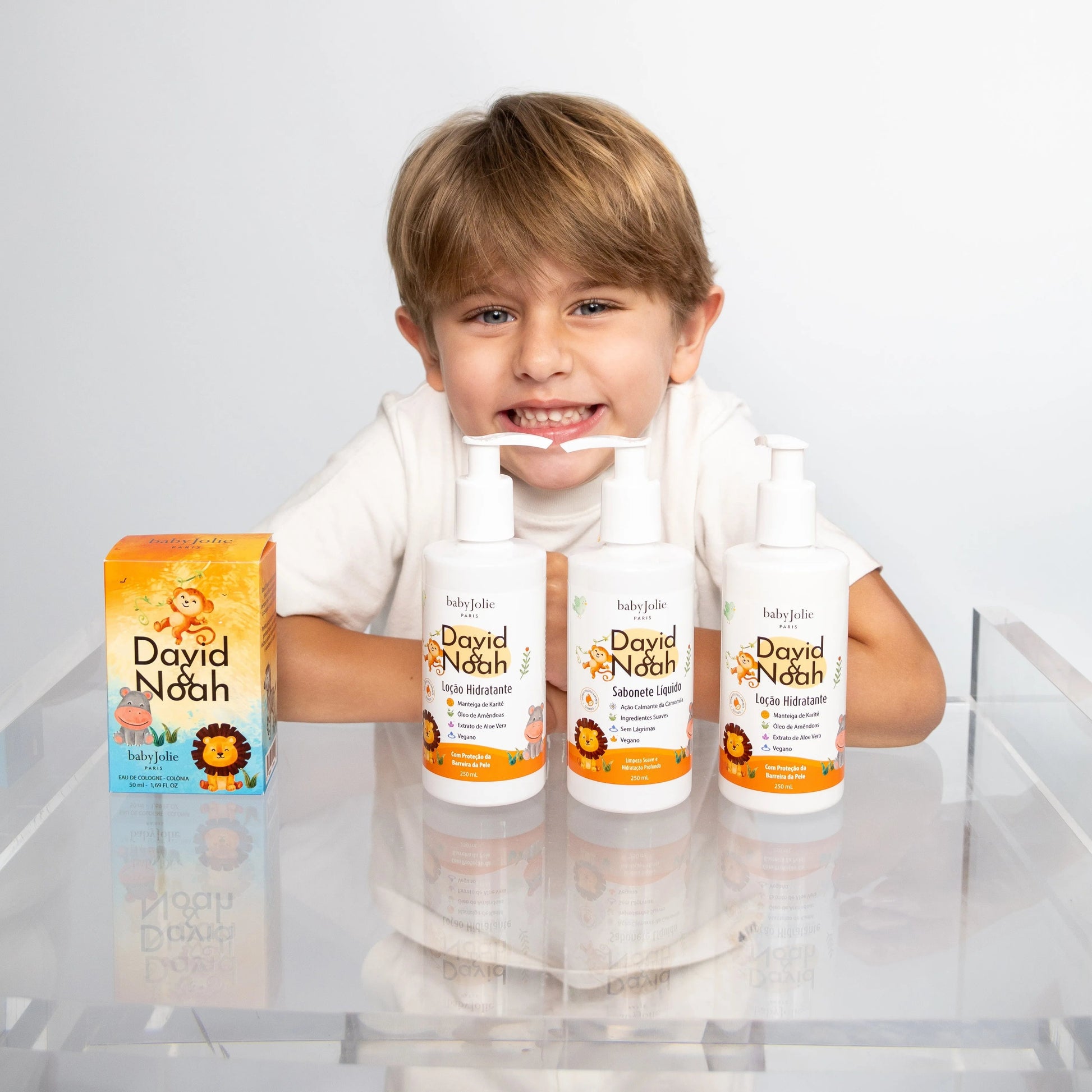 Smiling boy behind David & Noah baby skincare products, including a perfume box and three white pump bottles on a clear table.