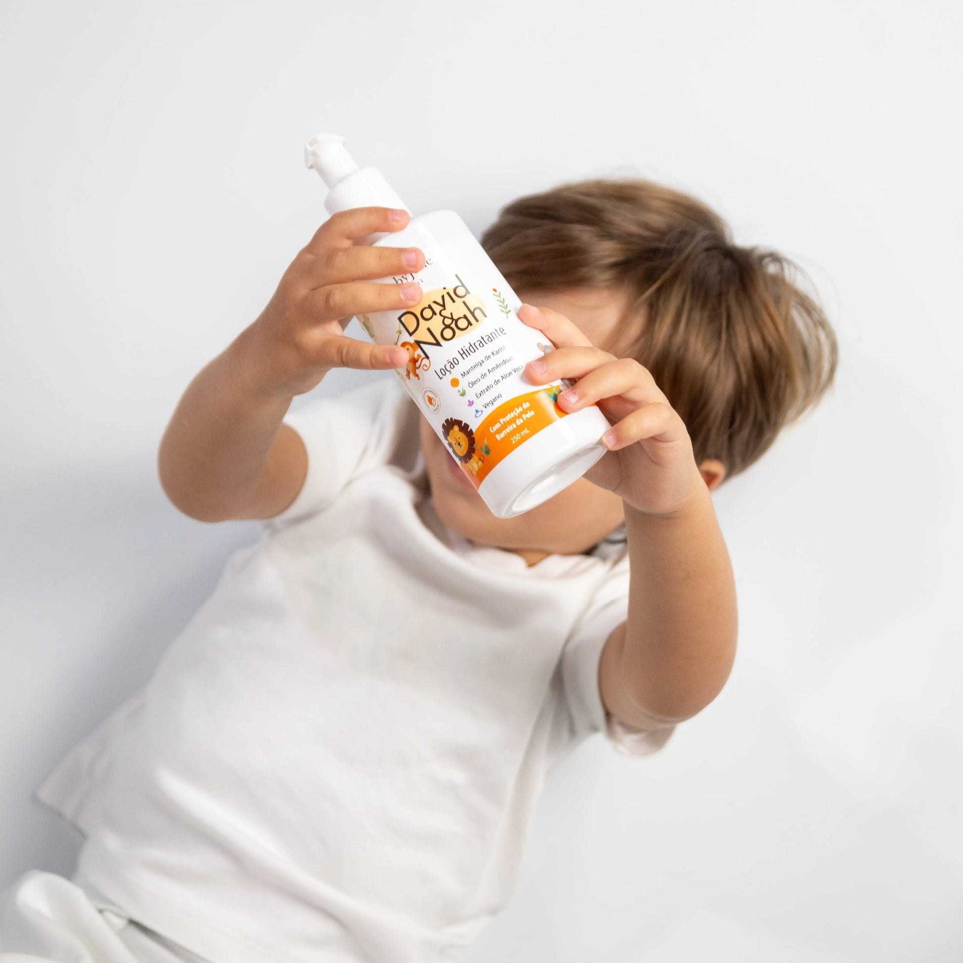 Toddler in a white shirt holding a white David & Noah moisturizing lotion bottle with a cute lion illustration.