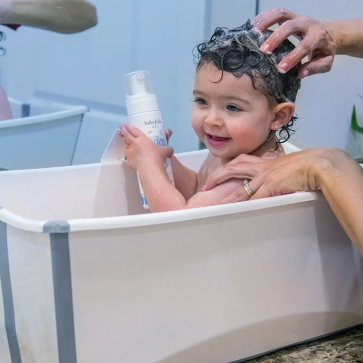 Smiling baby in a white collapsible tub holding a bottle of Baby Jolie foam shampoo while getting hair washed.