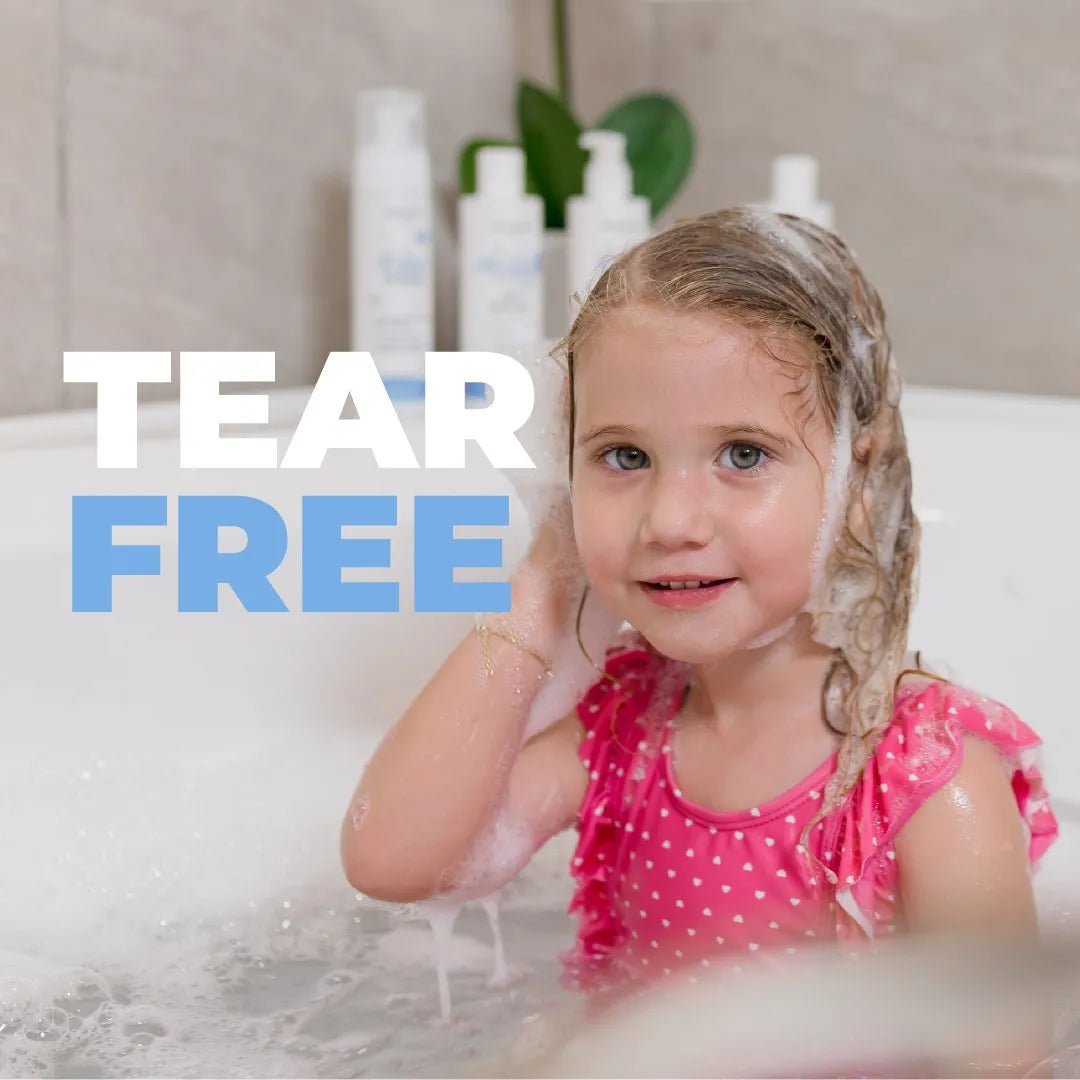 Young girl in a pink polka-dot swimsuit washing hair with tear-free shampoo in a bathtub with Baby Jolie product bottles.