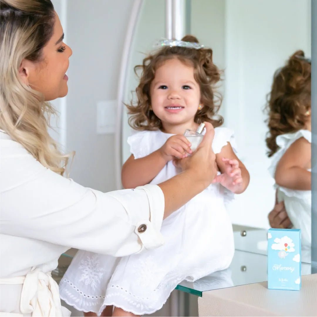 Mother applying Memory for Babies perfume to a smiling toddler in a white dress next to a light blue product box.